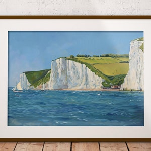 May include: A framed painting shows white cliffs, green hills, and a blue sea under a clear sky. A sailboat is on the water. The artwork is displayed on a wooden surface against a white brick wall.