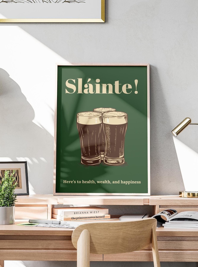 Może przedstawiać: Zielony plakat z tekstem "Sl&aacute;inte!" w kolorze kremowym. Pod tekstem przedstawiono trzy szklanki ciemnego piwa. Tekst "Here's to health, wealth, and happiness" znajduje się na dole plakatu.