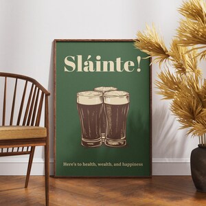 Może przedstawiać: Zielony plakat z tekstem "Sl&aacute;inte!" w kolorze białym i grafiką trzech szklanek ciemnego piwa. Tekst pod szklankami brzmi "Here's to health, wealth, and happiness."