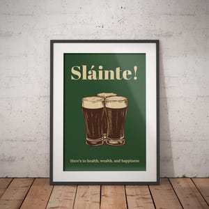 May include: A framed print with a green background and the text "Sl&aacute;inte!" in a yellow font. Three glasses of dark beer are arranged in a triangle shape. The text "Here's to health, wealth, and happiness" is below the glasses.