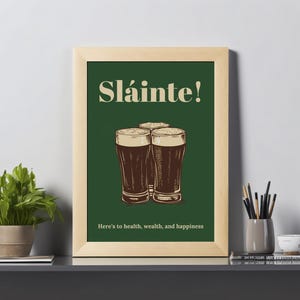Może przedstawiać: Wydruk w ramie z zielonym tłem i tekstem "Sl&aacute;inte!" w białym kolorze. Pod tekstem przedstawiono trzy szklanki ciemnego piwa. Tekst "Here's to health, wealth, and happiness" znajduje się na dole wydruku.