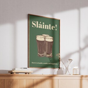 Może przedstawiać: Zielony plakat z napisem "Sl&aacute;inte!" w kolorze białym i rysunkiem trzech szklanek ciemnego piwa. Tekst pod szklankami brzmi "Here's to health, wealth, and happiness."
