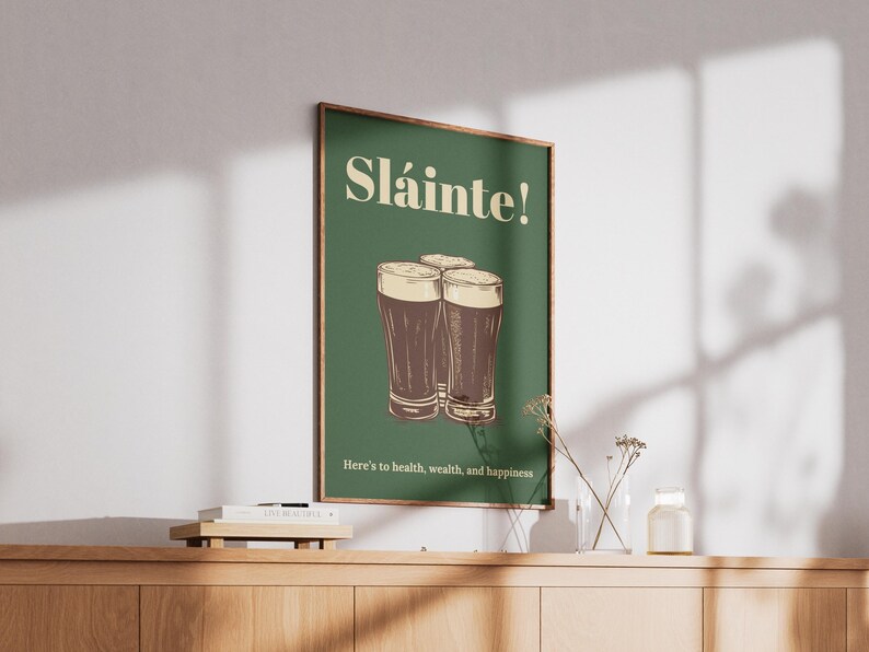 Może przedstawiać: Zielony plakat z napisem "Sl&aacute;inte!" w kolorze białym i rysunkiem trzech szklanek ciemnego piwa. Tekst pod szklankami brzmi "Here's to health, wealth, and happiness."