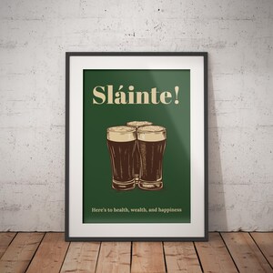 Może przedstawiać: Wydruk w ramie z zielonym tłem i tekstem "Sl&aacute;inte!" w kolorze ż&oacute;łtym. Trzy szklanki ciemnego piwa ułożone są w kształcie tr&oacute;jkąta. Tekst "Here's to health, wealth, and happiness" znajduje się na dole wydruku.