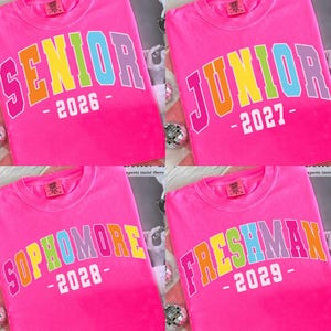 Puede incluir: Cuatro camisetas rosas brillantes con texto colorido. Cada camiseta muestra un año de clase diferente: "SENIOR 2026", "JUNIOR 2027", "SOPHOMORE 2028" y "FRESHMAN 2029". El texto está en degradado arcoíris.