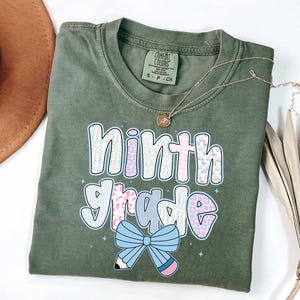 Pu&ograve; includere: T-shirt verde oliva con la scritta "ninth grade" in un design colorato e fantasia. Il design include un fiocco azzurro e una matita. Sono visibili anche una collana d'oro e un cappello marrone.