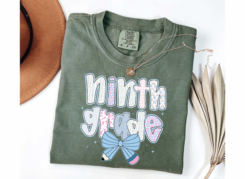 Pu&ograve; includere: T-shirt verde oliva con la scritta "ninth grade" in un design colorato e fantasia. Il design include un fiocco azzurro e una matita. Sono visibili anche una collana d'oro e un cappello marrone.