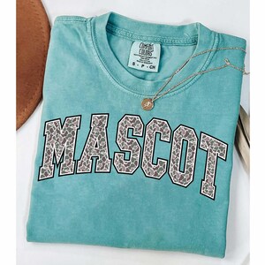 May include: A turquoise Comfort Colors t-shirt with the word "MASCOT" in a gray and white animal print design. A gold necklace with a pendant rests on the shirt. The shirt is folded on a white surface.