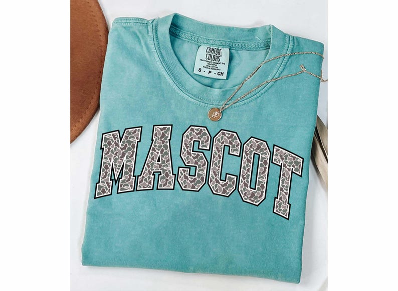 May include: A turquoise Comfort Colors t-shirt with the word "MASCOT" in a gray and white animal print design. A gold necklace with a pendant rests on the shirt. The shirt is folded on a white surface.