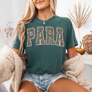 May include: A teal t-shirt featuring the word "PARA" in bold, leopard print lettering. The shirt is paired with denim shorts and a cream cardigan. The overall style is casual and relaxed.