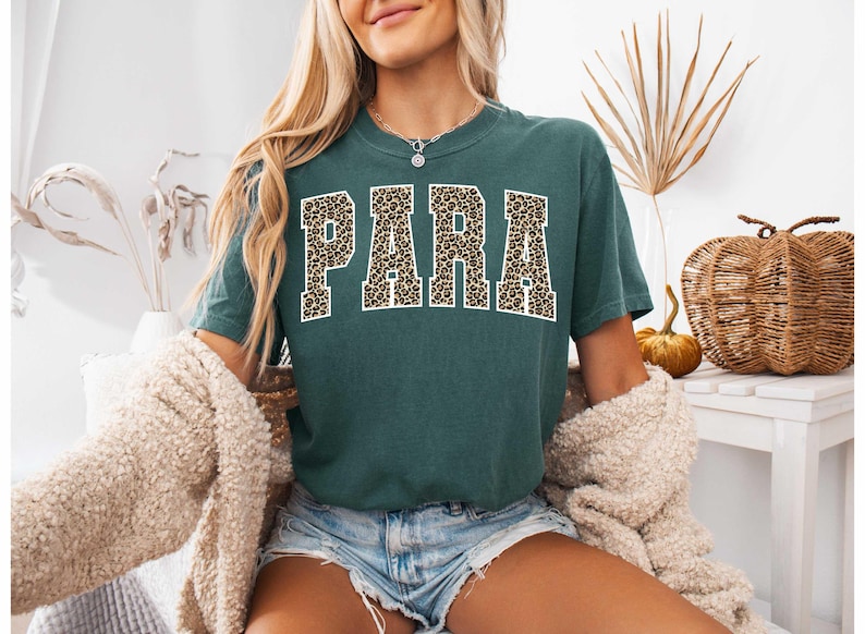 May include: A teal t-shirt featuring the word "PARA" in bold, leopard print lettering. The shirt is paired with denim shorts and a cream cardigan. The overall style is casual and relaxed.