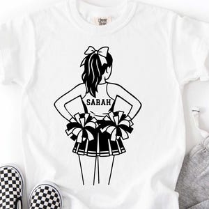 May include: White t-shirt featuring a black and white illustration of a cheerleader with pom-poms. The cheerleader's name, "SARAH," is printed on the back of her top. The shirt is paired with black and white checkered shoes.