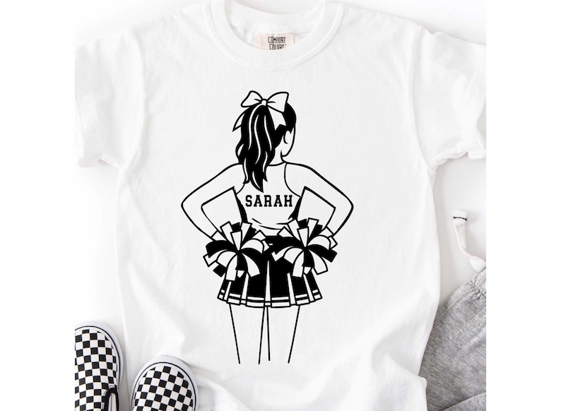 May include: White t-shirt featuring a black and white illustration of a cheerleader with pom-poms. The cheerleader's name, "SARAH," is printed on the back of her top. The shirt is paired with black and white checkered shoes.