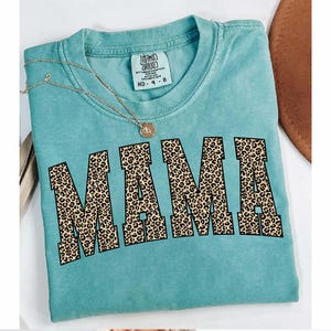 May include: A turquoise t-shirt with the word "MAMA" in large, leopard print letters across the front. A gold necklace with a pendant rests on the shirt. The shirt has a tag that reads "MIND CHILD" and "MD 9-8".