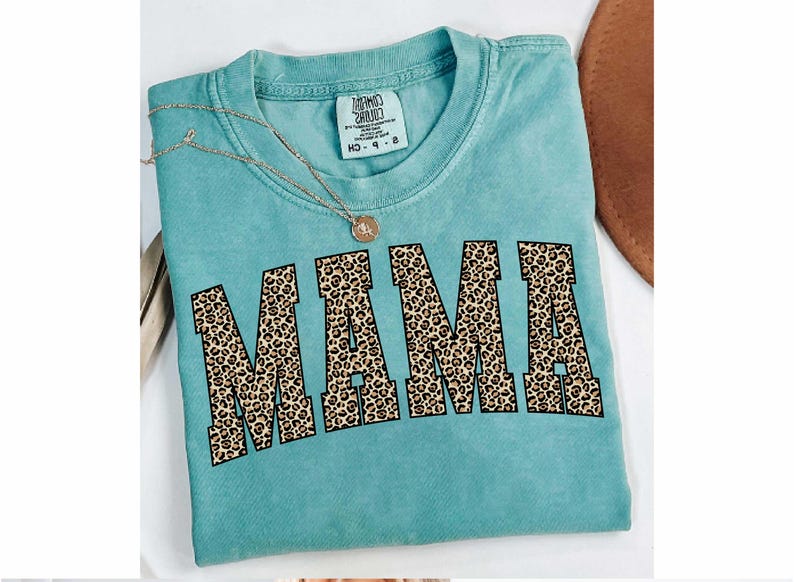 May include: A turquoise t-shirt with the word "MAMA" in large, leopard print letters across the front. A gold necklace with a pendant rests on the shirt. The shirt has a tag that reads "MIND CHILD" and "MD 9-8".