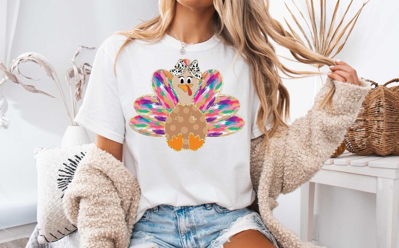 Op de afbeelding: Een wit T-shirt met een kleurrijk kalkoenontwerp. De kalkoen heeft een bruin lichaam met witte stippen, een roze kop en een zwart-witte strik. De veren van de kalkoen zijn een mix van roze, paars, blauw en groen.