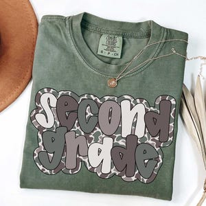 May include: Olive green t-shirt with the words "Second Grade" in a layered design. The text is outlined in white and gray, with a camouflage pattern. A gold necklace and a brown hat are visible.