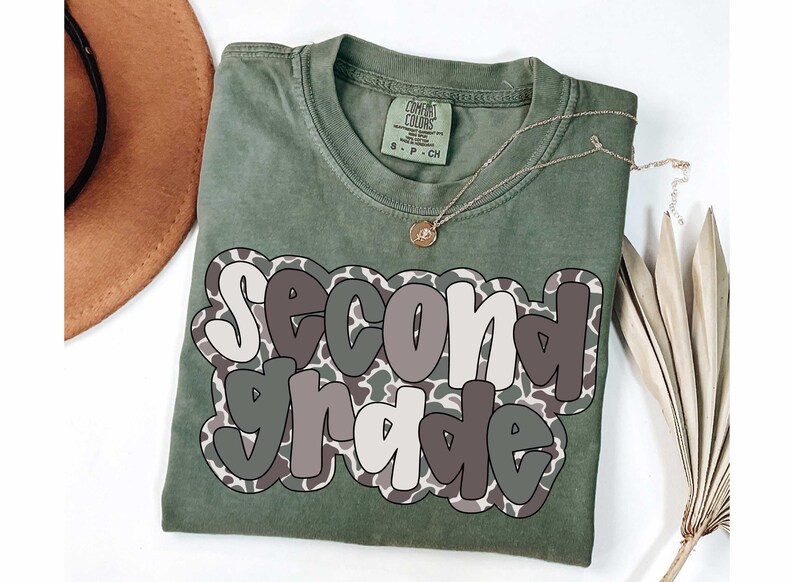 May include: Olive green t-shirt with the words "Second Grade" in a layered design. The text is outlined in white and gray, with a camouflage pattern. A gold necklace and a brown hat are visible.