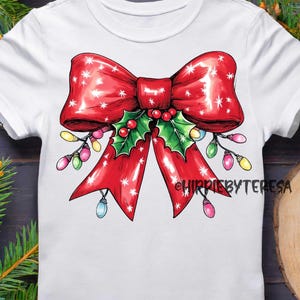 May include: White t-shirt featuring a large red Christmas bow with white star accents, holly, and colorful string lights. The text "HIPPIEBYTERESA" is printed on the right side of the bow. Festive apparel.