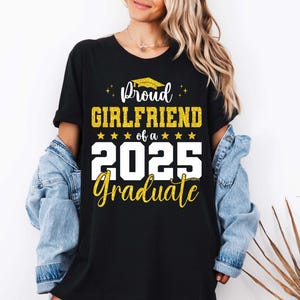 K&ouml;nnte beinhalten: Schwarzes T-Shirt mit wei&szlig;em Text, der "Proud Girlfriend of a 2025 Graduate" lautet. Der Text ist mit goldenem Glitzer und einer goldenen Abschluss-Kappe verziert.