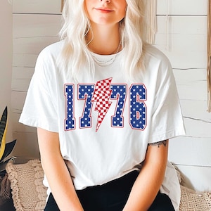 May include: White t-shirt with a red, white, and blue graphic of the number 1776 with a lightning bolt in the middle. The numbers and lightning bolt are outlined in blue.
