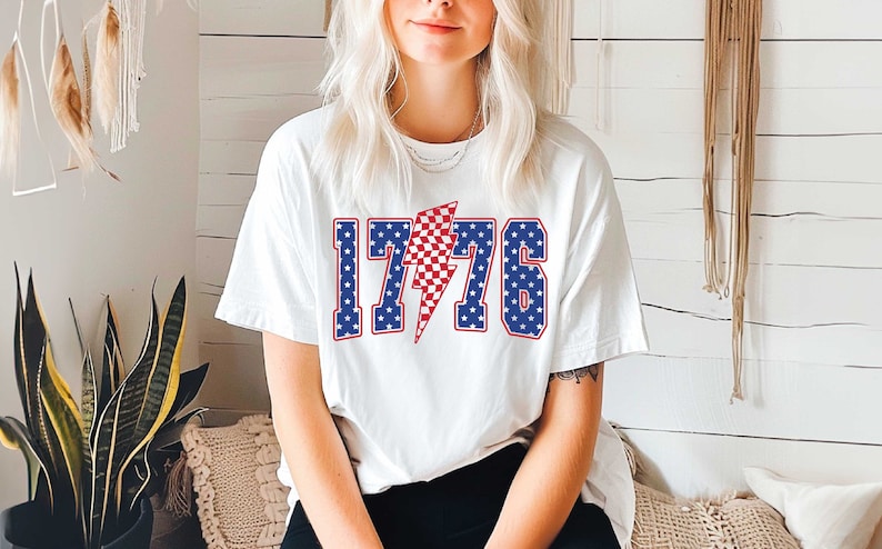 May include: White t-shirt with a red, white, and blue graphic of the number 1776 with a lightning bolt in the middle. The numbers and lightning bolt are outlined in blue.