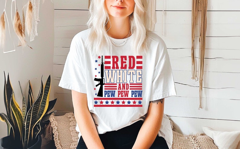 May include: White t-shirt with a red, white, and blue graphic design. The graphic features a stylized rifle with the words "RED WHITE AND PEW PEW PEW" and stars.