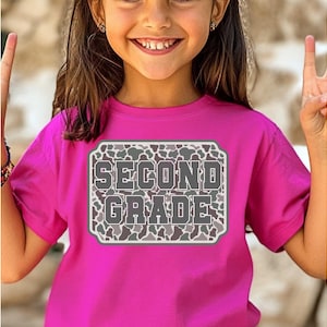 May include: A bright pink t-shirt with the words "SECOND GRADE" in a gray, camouflage-patterned design. The text is inside a white-bordered rectangle. The shirt is being worn by a young person.