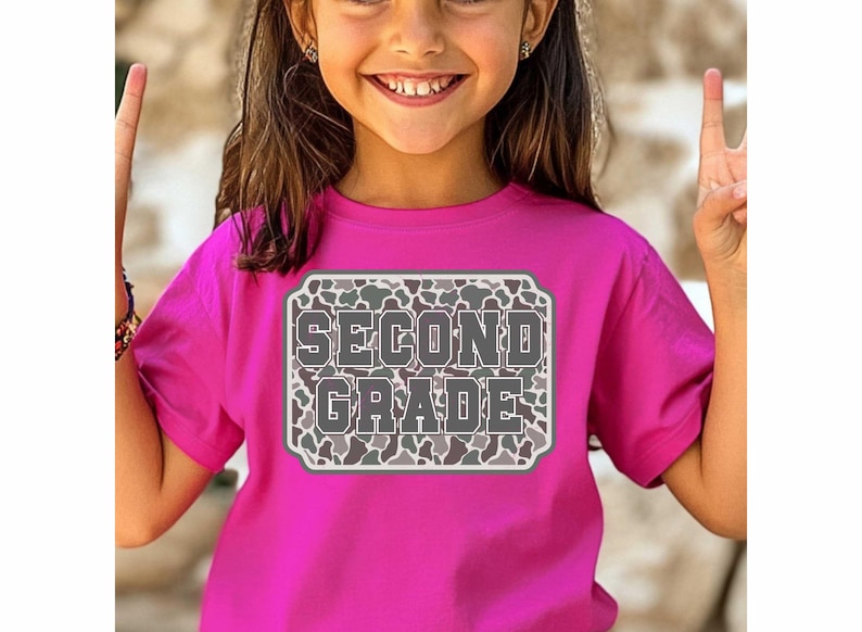 May include: A bright pink t-shirt with the words "SECOND GRADE" in a gray, camouflage-patterned design. The text is inside a white-bordered rectangle. The shirt is being worn by a young person.