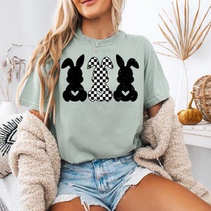 Puede incluir: Una camiseta verde claro con un gr&aacute;fico de tres conejos negros y blancos. Los conejos est&aacute;n en fila, con el conejo del medio con un patr&oacute;n a cuadros. Cada conejo tiene un peque&ntilde;o coraz&oacute;n en el pecho.