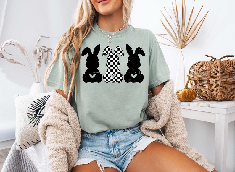 Puede incluir: Una camiseta verde claro con un gr&aacute;fico de tres conejos negros y blancos. Los conejos est&aacute;n en fila, con el conejo del medio con un patr&oacute;n a cuadros. Cada conejo tiene un peque&ntilde;o coraz&oacute;n en el pecho.