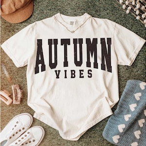 May include: Cream-colored t-shirt with the words "AUTUMN VIBES" in bold, black, distressed lettering. The shirt is styled with white sneakers, a brown corduroy hat, and a blue blanket with white hearts. A silver necklace is visible.