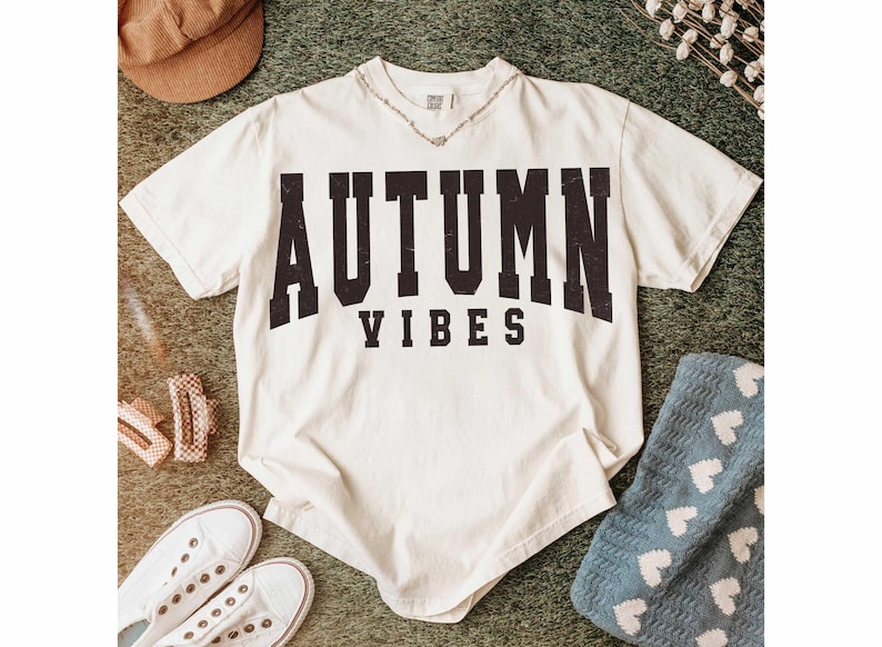 May include: Cream-colored t-shirt with the words "AUTUMN VIBES" in bold, black, distressed lettering. The shirt is styled with white sneakers, a brown corduroy hat, and a blue blanket with white hearts. A silver necklace is visible.