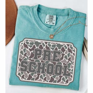 May include: A turquoise Comfort Colors t-shirt with a camouflage design and the words "PRE SCHOOL" in a gray, block letter font. A gold necklace with a pendant rests on the shirt. The shirt is folded on a white surface.