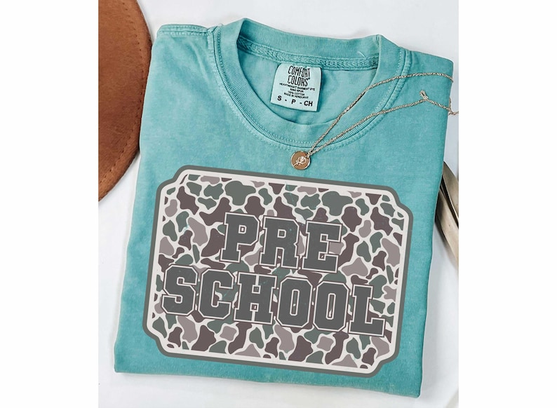 May include: A turquoise Comfort Colors t-shirt with a camouflage design and the words "PRE SCHOOL" in a gray, block letter font. A gold necklace with a pendant rests on the shirt. The shirt is folded on a white surface.
