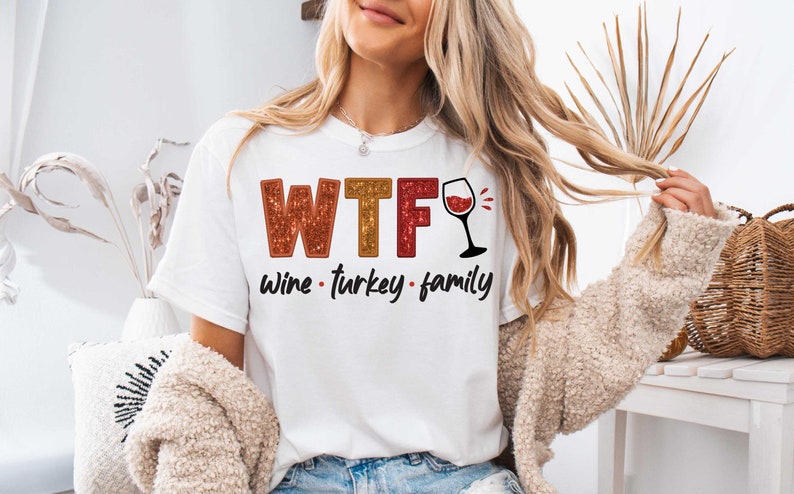 May include: White t-shirt with the text "WTF Wine - Turkey - Family" in red, orange, and yellow glitter lettering. A red wine glass with a red wine splash is next to the text.