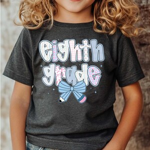 Pu&ograve; includere: T-shirt grigio scuro di "Eighth Grade" con un design colorato. Il testo "Eighth Grade" &egrave; in un font a bolle con accenti floreali e pastello. Un fiocco azzurro e una matita adornano la parte inferiore del testo.