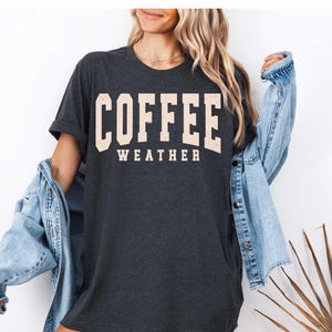 May include: Dark gray t-shirt with the words "COFFEE WEATHER" in a cream-colored, collegiate-style font. The shirt is worn with a light blue denim jacket.
