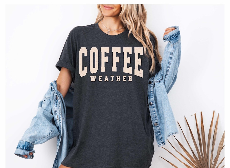 May include: Dark gray t-shirt with the words "COFFEE WEATHER" in a cream-colored, collegiate-style font. The shirt is worn with a light blue denim jacket.