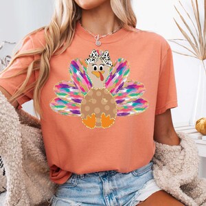 Op de afbeelding: Een perzikkleurig T-shirt met een kleurrijke kalkoenprint. De kalkoen heeft een bruin lichaam met witte stippen, een roze kop en een roze strik met witte stippen. De veren van de kalkoen zijn regenboogkleurig.
