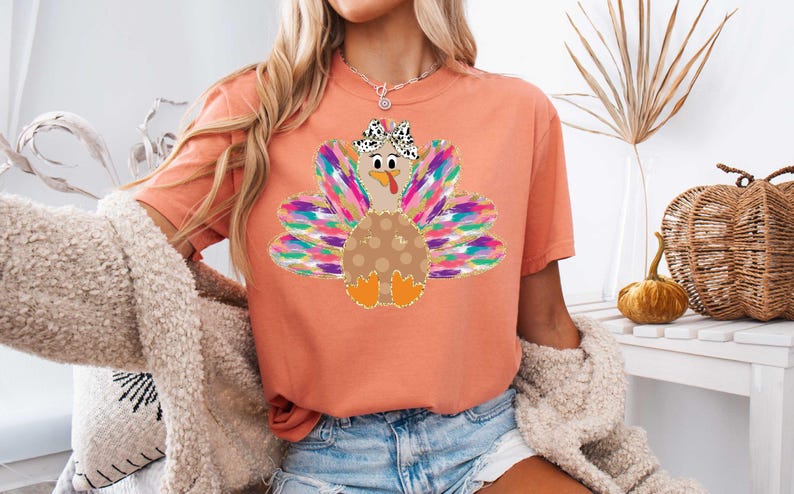 Op de afbeelding: Een perzikkleurig T-shirt met een kleurrijke kalkoenprint. De kalkoen heeft een bruin lichaam met witte stippen, een roze kop en een roze strik met witte stippen. De veren van de kalkoen zijn regenboogkleurig.