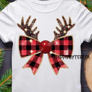 May include: White t-shirt featuring a festive design. The graphic includes reindeer antlers with a starry pattern, a red and black plaid bow, and a glittery red ball. The design is outlined in gold. The text "HIPPIEBYTERESA" is visible.