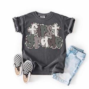 May include: A dark gray t-shirt with the words "first grade" in a bubble letter style with a camouflage pattern. The shirt is paired with light blue denim shorts and black and white checkered slip-on shoes.