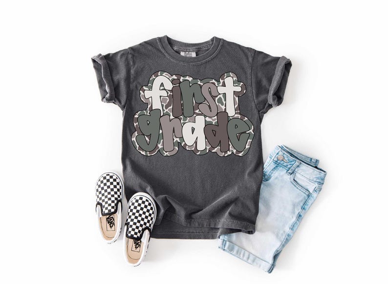 May include: A dark gray t-shirt with the words "first grade" in a bubble letter style with a camouflage pattern. The shirt is paired with light blue denim shorts and black and white checkered slip-on shoes.