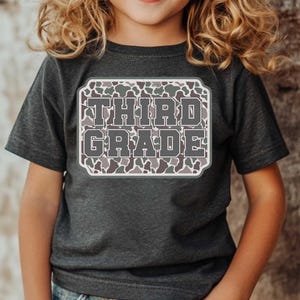 May include: A dark gray t-shirt featuring the words "THIRD GRADE" in a gray, block letter font. The text is set within a white-bordered, rectangular design with a camouflage pattern. The shirt is worn by a child.
