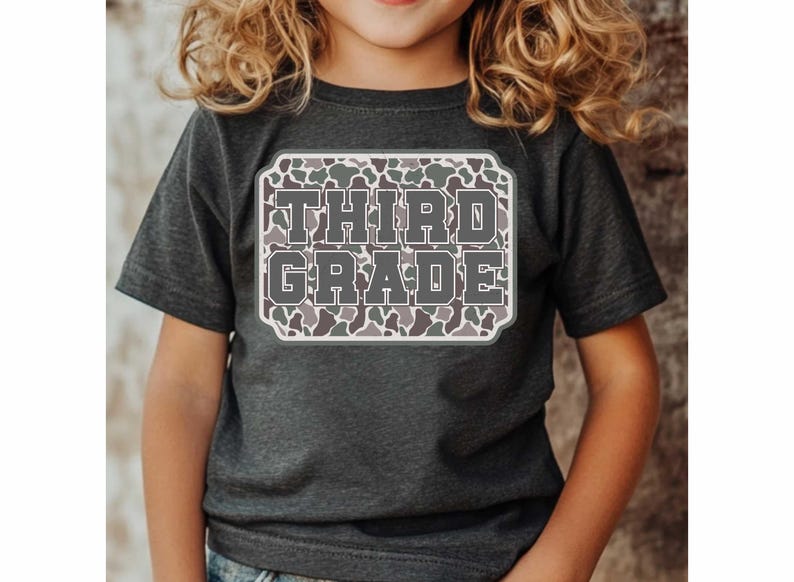 May include: A dark gray t-shirt featuring the words "THIRD GRADE" in a gray, block letter font. The text is set within a white-bordered, rectangular design with a camouflage pattern. The shirt is worn by a child.