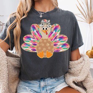 Op de afbeelding: Een donkergrijs T-shirt met een kleurrijke kalkoenprint. De kalkoen heeft een bruin lichaam met witte stippen, een roze kop en een zwart-witte strik. De veren van de kalkoen zijn een mix van roze, paars, blauw, groen en goud.