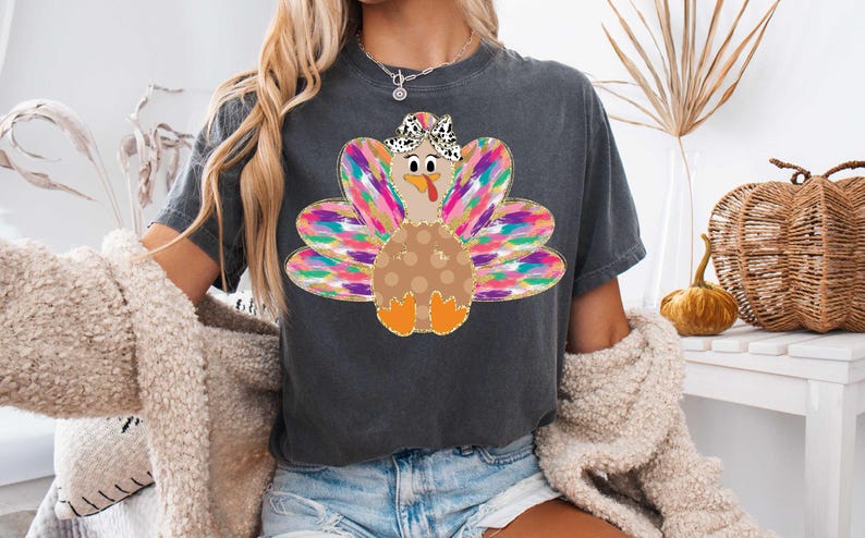 Op de afbeelding: Een donkergrijs T-shirt met een kleurrijke kalkoenprint. De kalkoen heeft een bruin lichaam met witte stippen, een roze kop en een zwart-witte strik. De veren van de kalkoen zijn een mix van roze, paars, blauw, groen en goud.