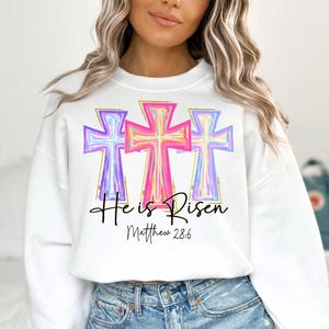Puede incluir: Sudadera blanca con un gr&aacute;fico de tres cruces coloridas y el texto "He is Risen" con la referencia b&iacute;blica "Mateo 28:6".