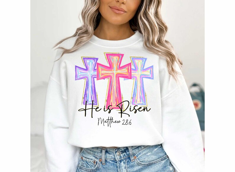 Puede incluir: Sudadera blanca con un gr&aacute;fico de tres cruces coloridas y el texto "He is Risen" con la referencia b&iacute;blica "Mateo 28:6".